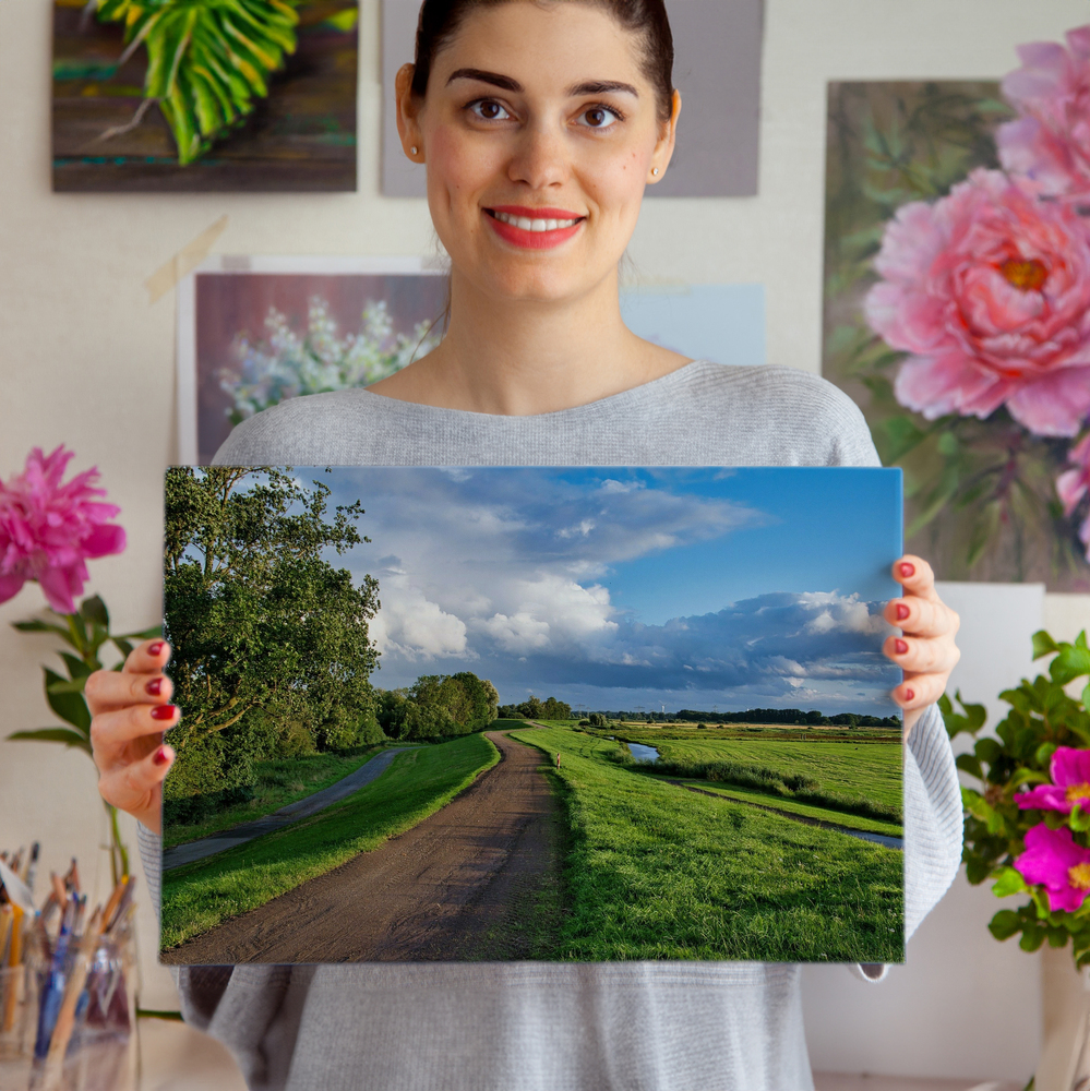 Randloze foto op plexiglas met landweg, groene weiden en wolkenlucht, voor een wand met bloemenkunst