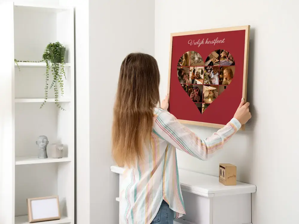 Kerstfotocollage in hartvorm op donkerrood met kerstgroet, in een lichte houten lijst aan een witte muur