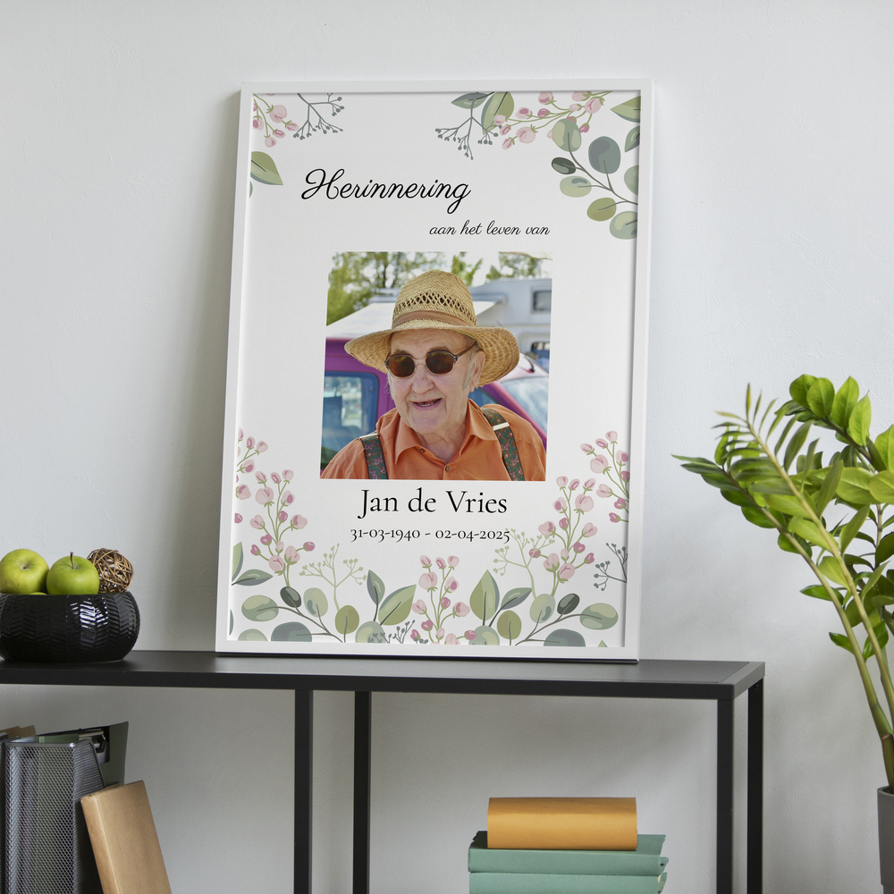 Herdenkingsposter met portret en bloemen in een witte lijst, op een zwarte kast naast een groene plant