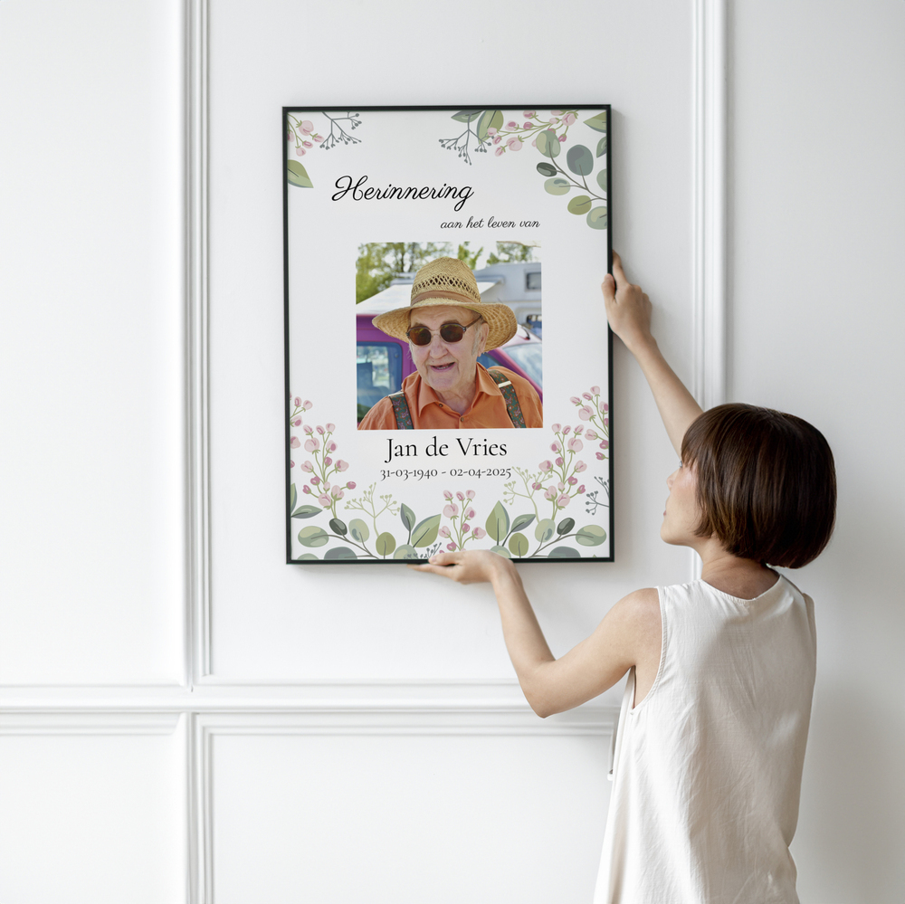 Rouwfoto met naam, datums en bloemenrand in een zwarte lijst aan een witte wand met sierlijsten